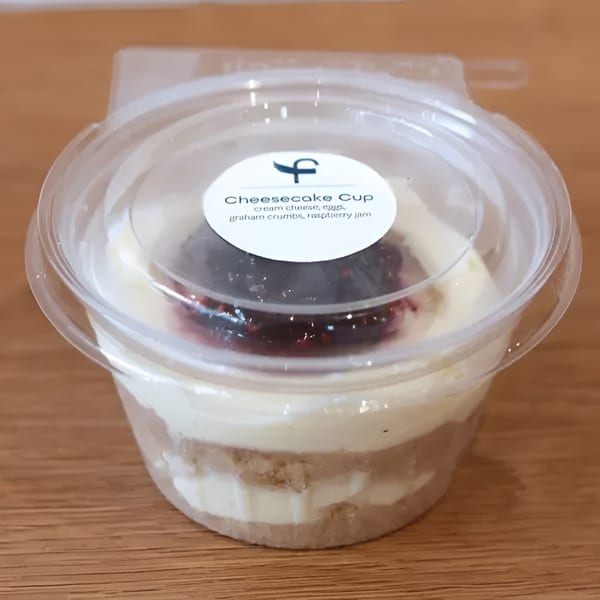 A clear plastic cup of cheesecake with raspberry jam on top, sitting on a wooden surface.