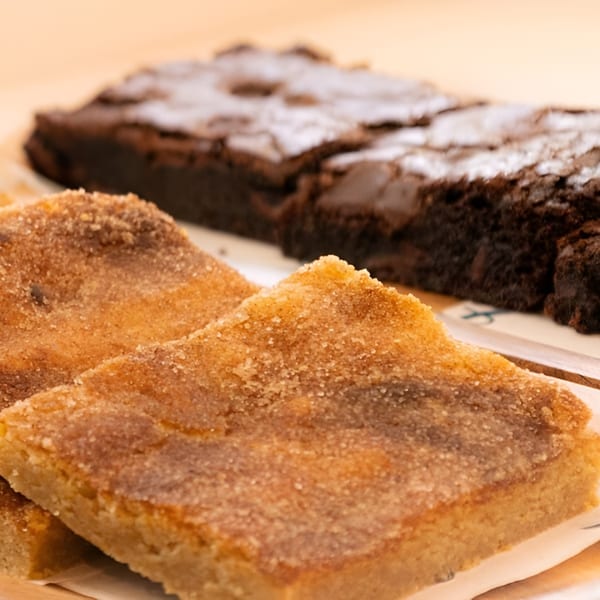 Close-up of two sugar-dusted blondies in the foreground, with blurred brownies in the background.