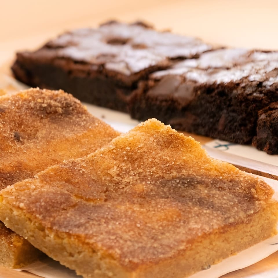Close-up of two sugar-dusted blondies in the foreground, with blurred brownies in the background.