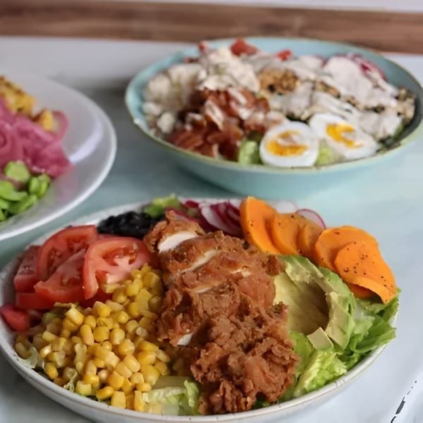 Three vibrant salads, one with fried chicken, corn, tomatoes, avocado, and sweet potato slices.