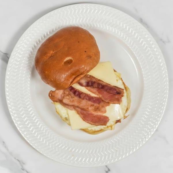 Top-down view of a breakfast sandwich with bacon, cheese, and egg on a white plate.