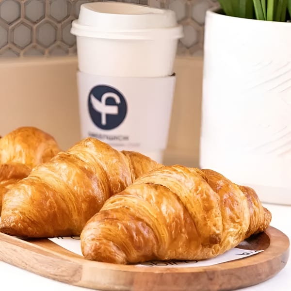 Golden-brown croissants on a wooden tray with a branded coffee cup and plant in the background.