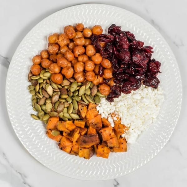 A white plate holds sections of chickpeas, dried cranberries, cottage cheese, roasted sweet potatoes, and pumpkin seeds.