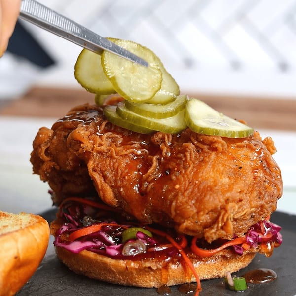 A chef uses tweezers to place pickles on a fried chicken sandwich with slaw and sauce.