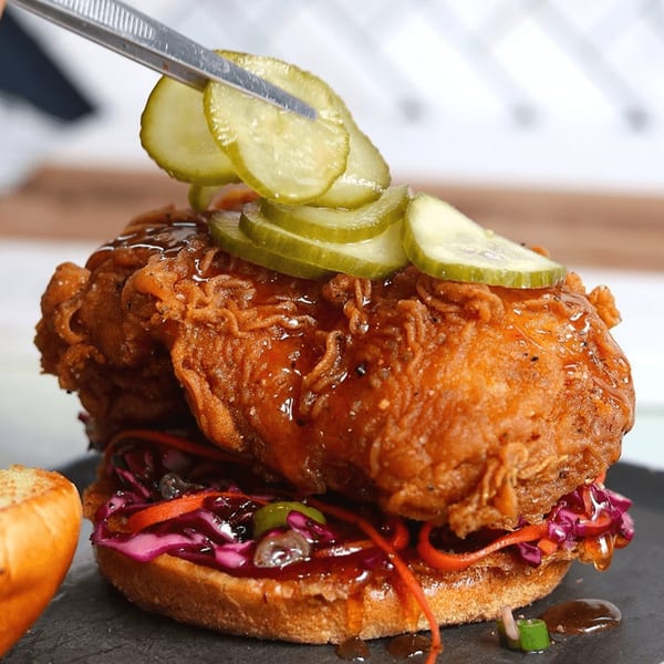 A chef uses tweezers to place pickles on a crispy fried chicken sandwich with slaw and sauce.
