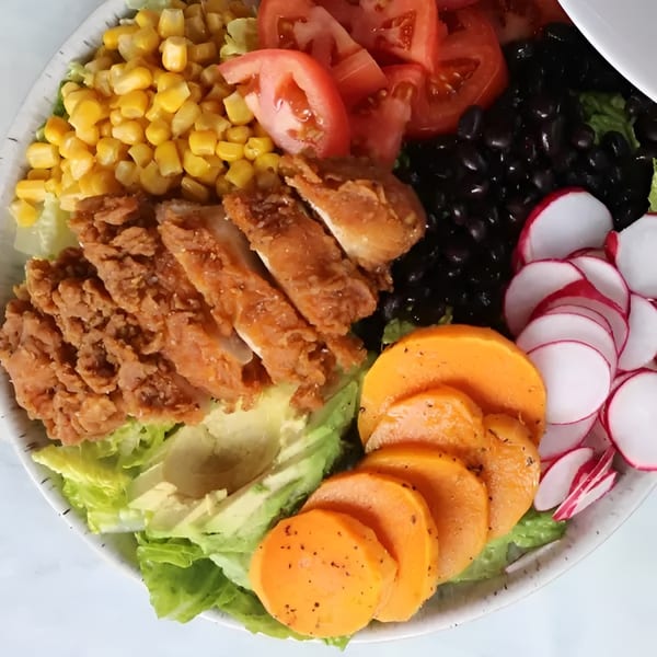 A vibrant salad bowl with fried chicken, corn, tomatoes, black beans, radishes, avocado, and sweet potato slices.