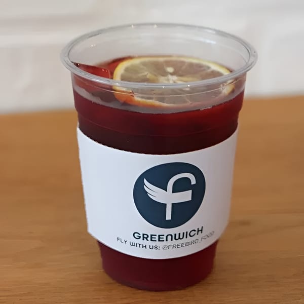 A Greenwich branded cup filled with a dark red beverage and lemon slices on a wooden surface.