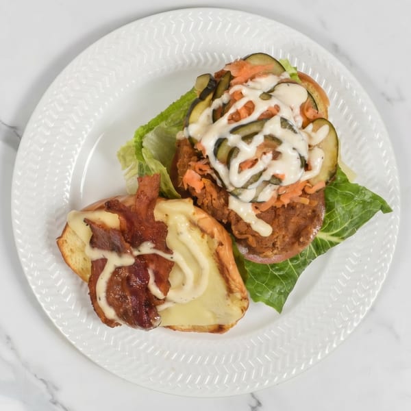 Open-faced sandwich with bacon, cheese, lettuce, pickles, shredded carrots, and white sauce on a white plate.