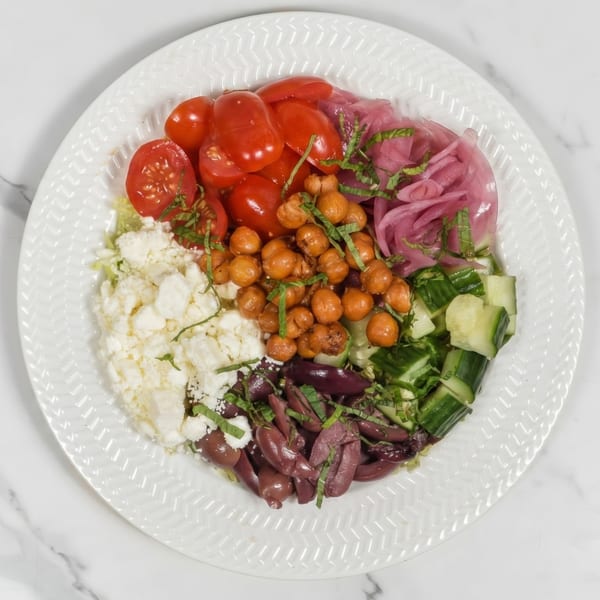 A colorful salad with tomatoes, pickled onions, chickpeas, feta, cucumbers, and olives on a white plate.