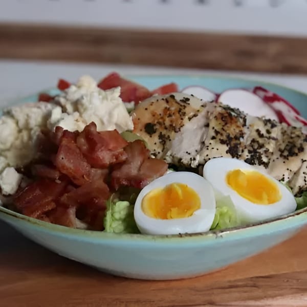 A vibrant salad with chicken, bacon, hard-boiled eggs, feta, and tomatoes in a blue bowl.