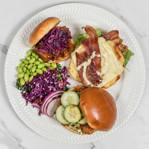 Three sliders with various toppings, edamame, and red cabbage salad on a white plate.