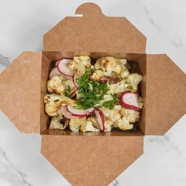 Roasted cauliflower and sliced radishes topped with green onions in a brown takeout box.