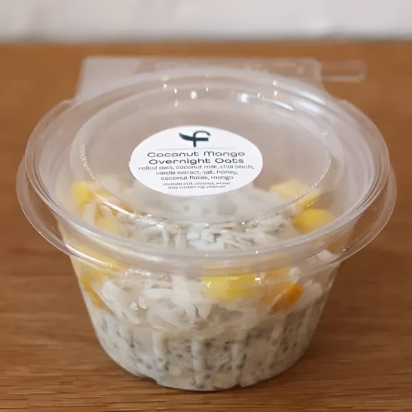 Coconut Mango Overnight Oats in a clear plastic container with a label on a wooden surface.