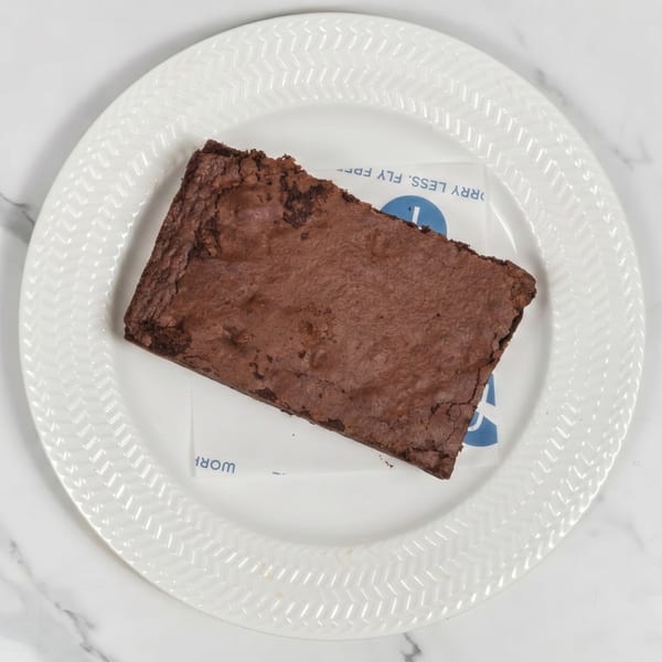 A rectangular chocolate brownie sits on a white plate with a patterned rim, on a marble surface.