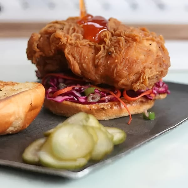 Crispy fried chicken sandwich with red cabbage slaw, drizzled with hot sauce, served with pickles.