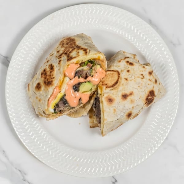 Two halves of a burrito filled with meat, avocado, and sauce, on a white plate.